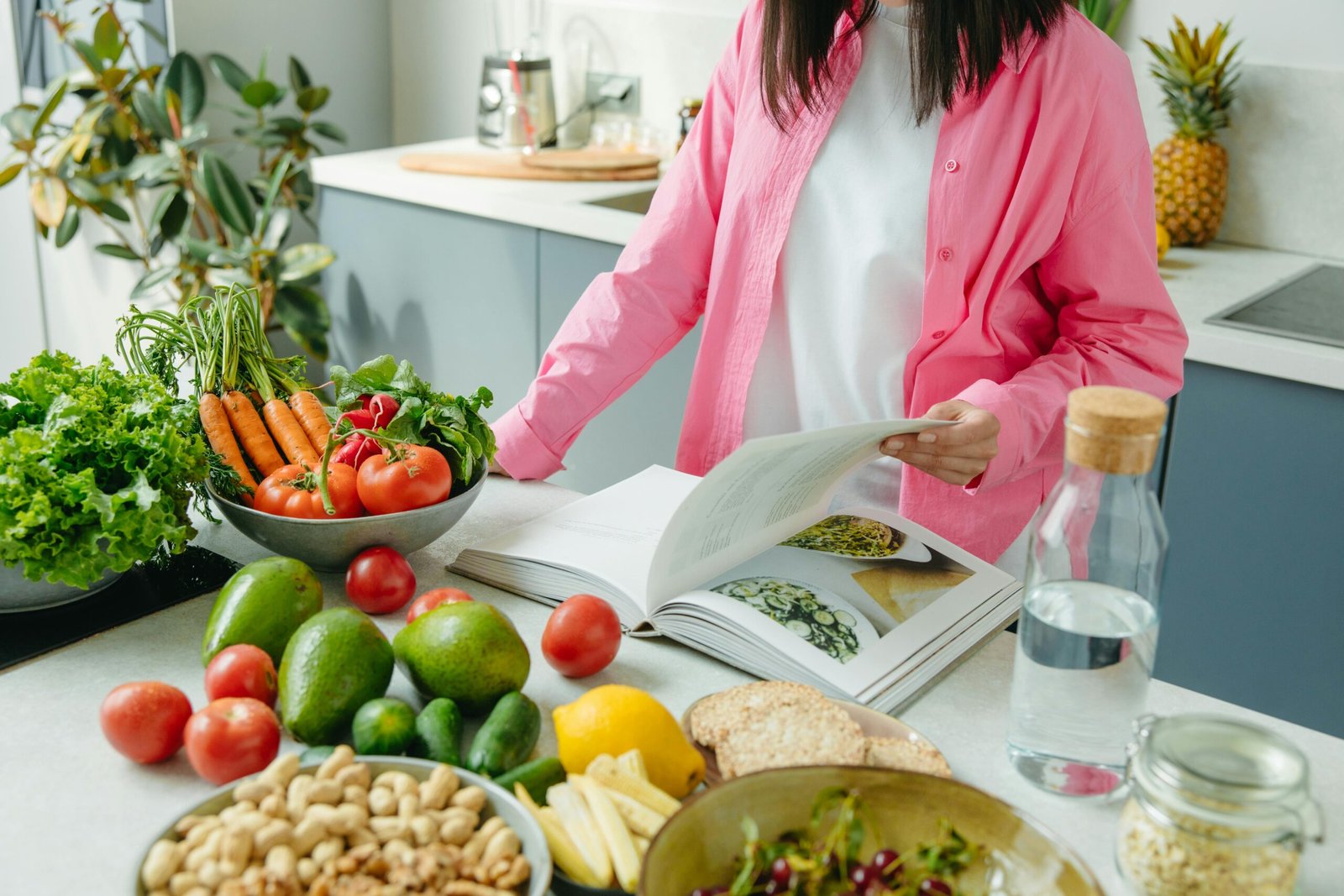 A person browsing a cookbook surrounded by fresh vegetables in a bright kitchen setting.